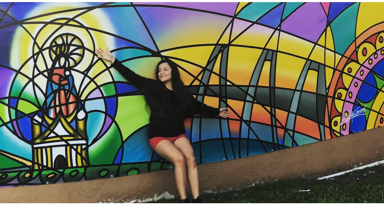 Lucía Gonmi artista de GonmiArt frente a un mural colorido de arte figurativo y devocional con diseño geométrico, inspiración espiritual y estilo único | Lucía Gonmi artist of GonmiArt in front of a colorful figurative and devotional mural with geometric design, spiritual inspiration, and unique style.